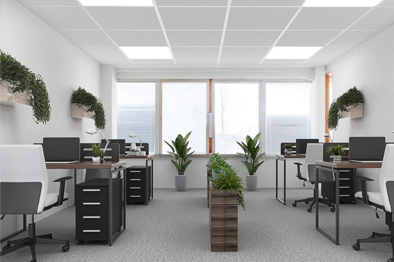 Moderni toimisto, jossa on kuusi työpistettä, mustat tuolit, tietokoneet ja vihreitä kasveja pöydillä ja hyllyillä. Suuret ikkunat antavat luonnonvaloa. Tilassa on neutraali värimaailma.