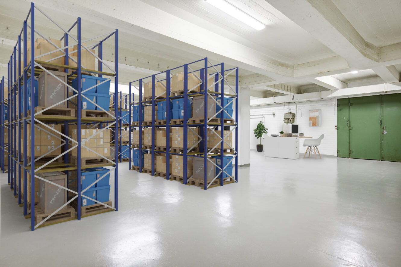 Varaston sisätilat, joissa on metallihyllyjä, joissa on sinisiä ja pahvisia säilytysastioita, ja toimistohuone, jossa on työpöytä ja tuoleja, taustalla vihreiden pariovien lähellä.