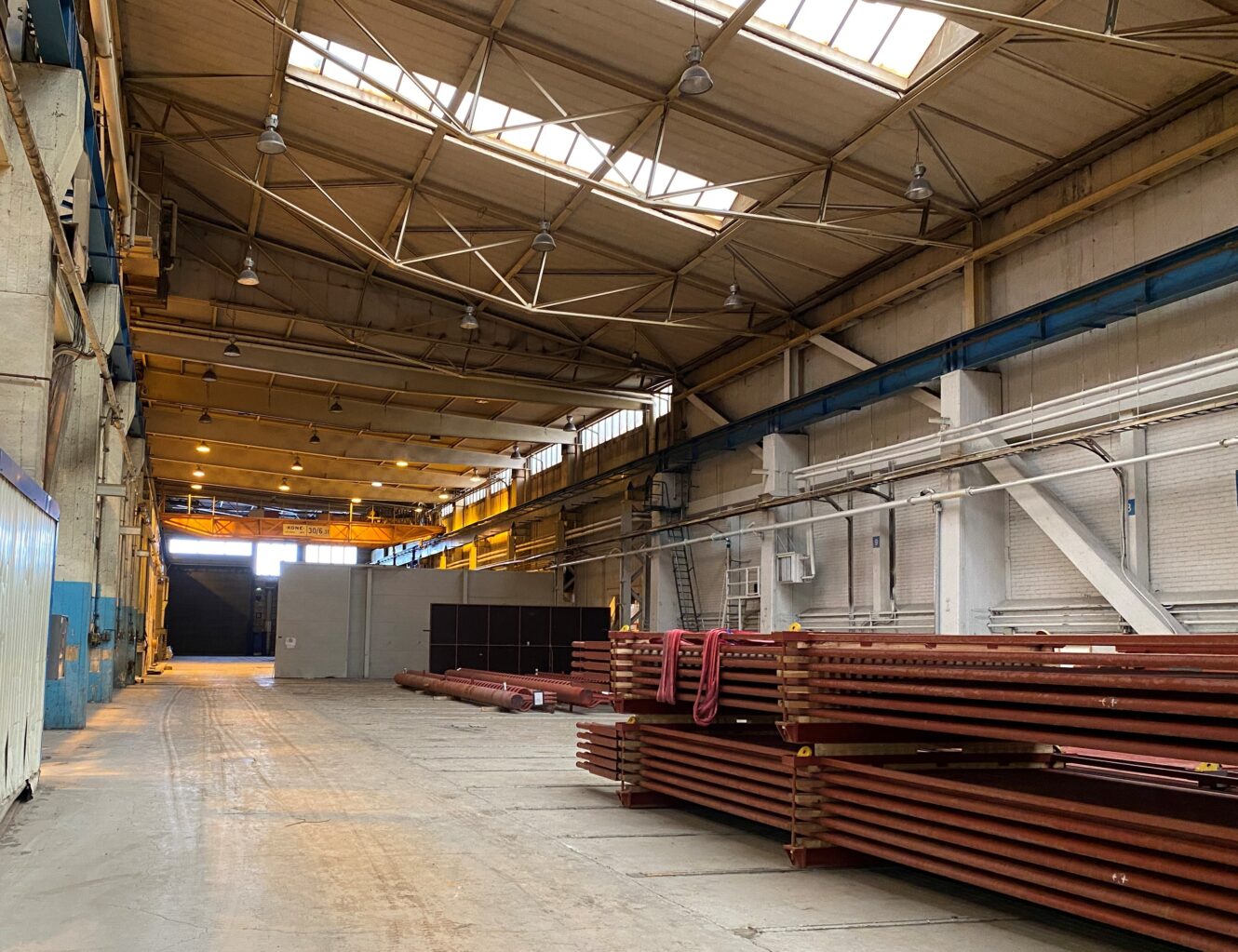 Suuren teollisuusvaraston sisätila, jossa on korkeat katot, pinotut metallipalkit, ylävalot ja avoin tila, joka johtaa suurelle ovelle.