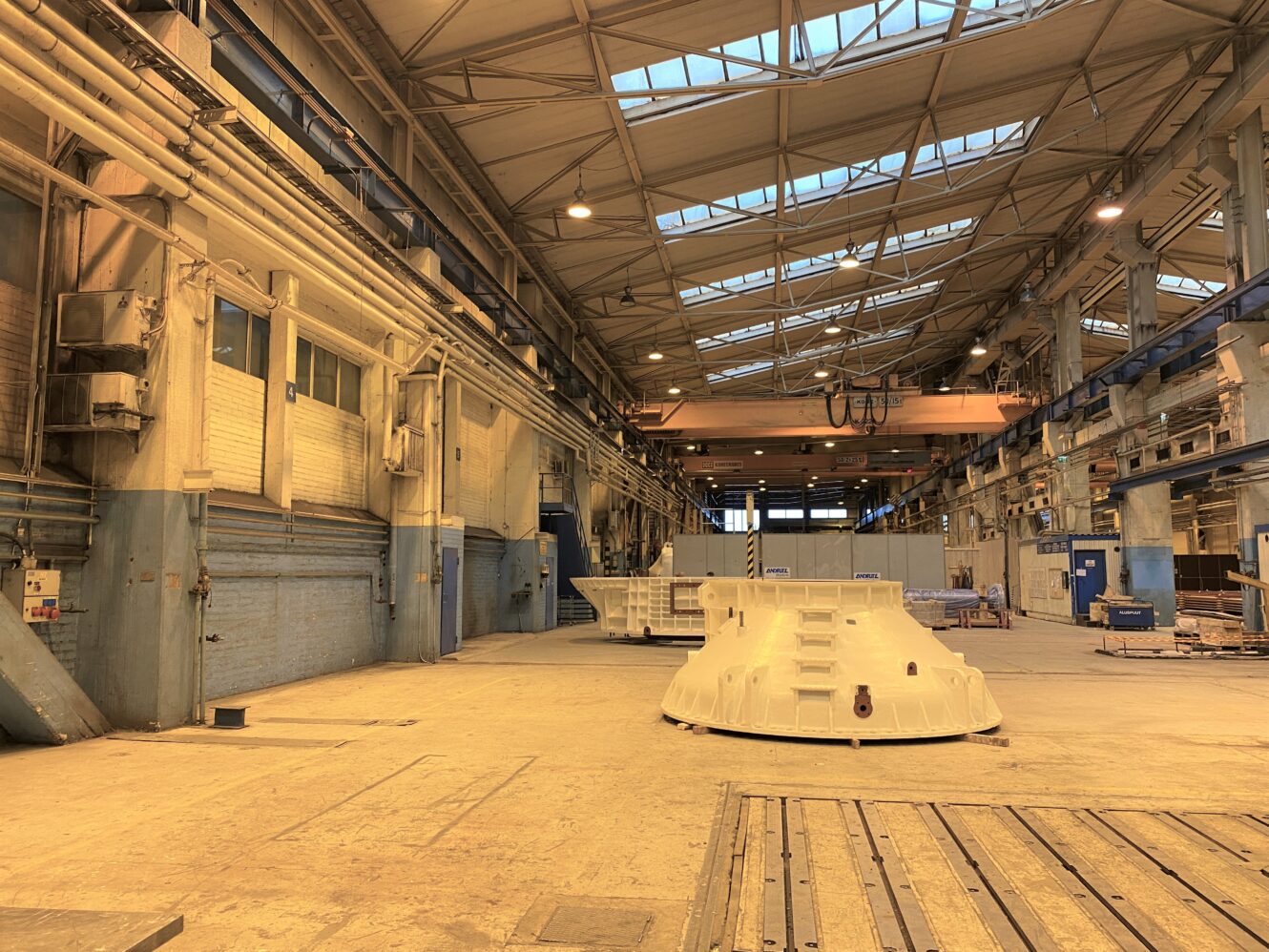 Suuri teollisuusvaraston sisätila, jossa on korkeat katot, kattoikkunat, nosturit ja lattialle sijoitetut koneenosat.