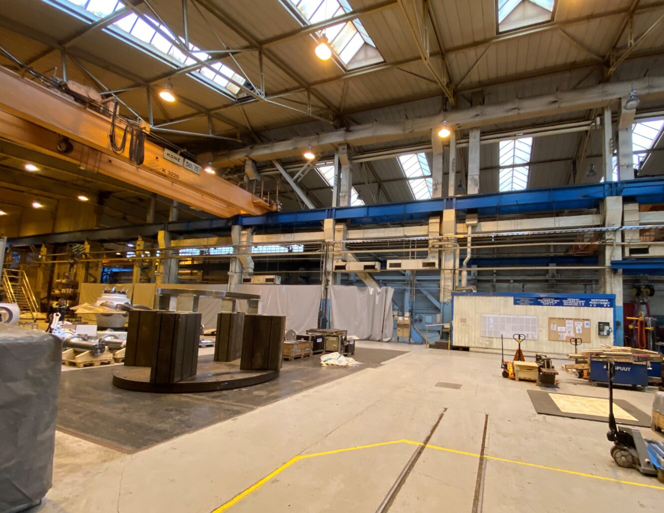 Tilava teollisuusvaraston sisätila, jossa on nosturi, suuria koneita, hajallaan olevia laitteita ja korkeita ikkunoita, jotka päästävät luonnonvaloa sisään.