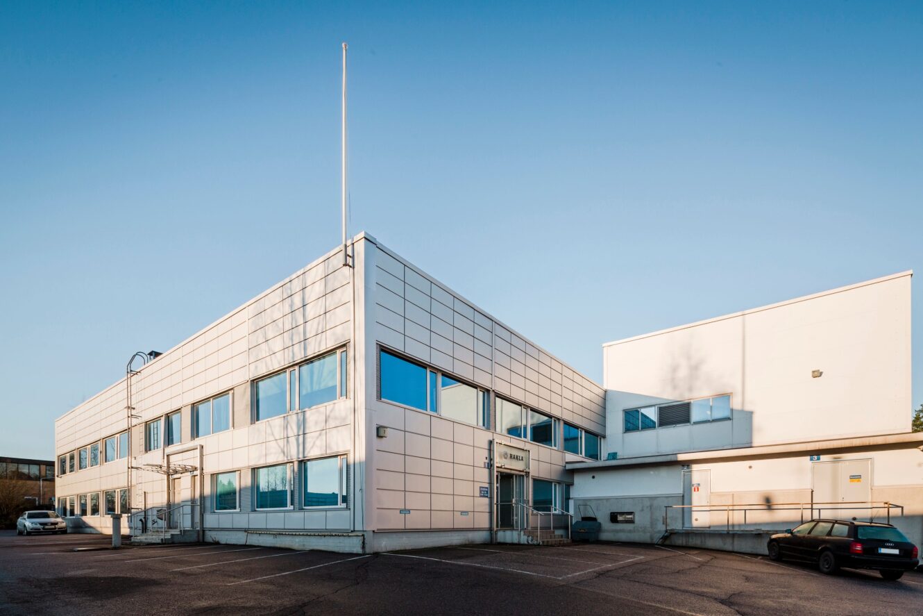 Moderni, valkoinen, kaksikerroksinen toimistorakennus, jossa on sinisävyiset ikkunat, tyhjä lipputanko ja pysäköity auto etualalla kirkkaalla taivaalla.