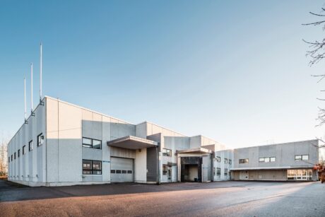 Suuri, moderni teollisuusrakennus, jossa on valkoiset ja harmaat ulkoseinät, leveät ikkunat ja useita autotallin ovia, kirkkaan sinisen taivaan alla.