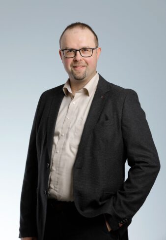 Lyhythiuksinen, silmälasipäinen ja parrakas mies, jolla on tumma bleiseri vaalean paidan päällä, seisoo ja hymyilee vaaleanharmaata taustaa vasten.