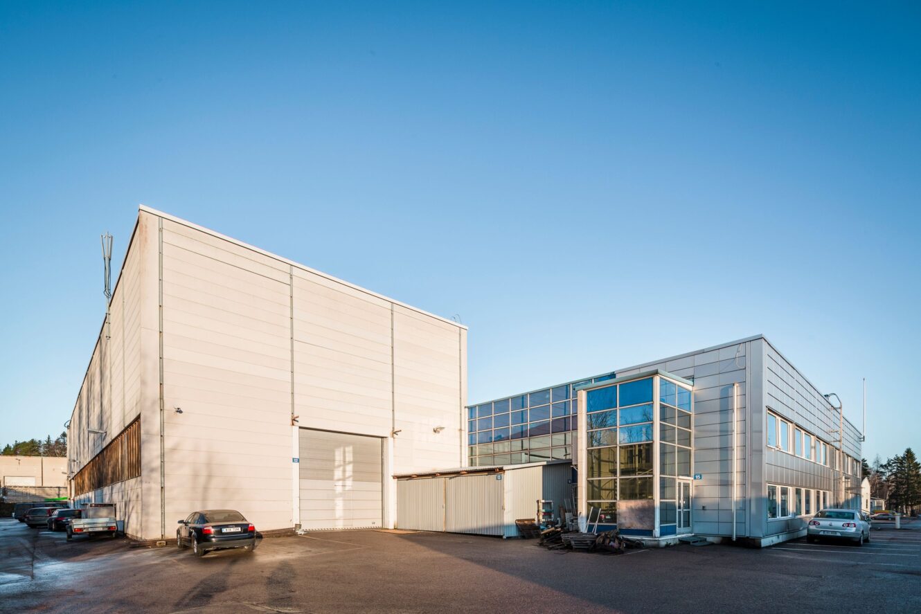 Moderni teollisuusrakennus, jossa on valkoiset ulkopaneelit, suuret ikkunat ja pysäköityjä autoja edessä kirkkaana, aurinkoisena päivänä.