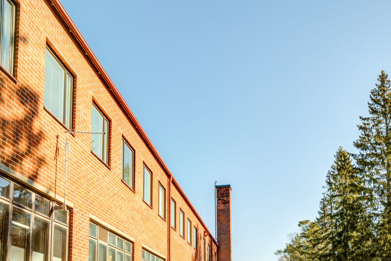 Punainen tiilirakennus, jossa on useita ikkunoita ja korkea savupiippu kirkkaalla sinisellä taivaalla, ja oikealla puolella näkyy puita.