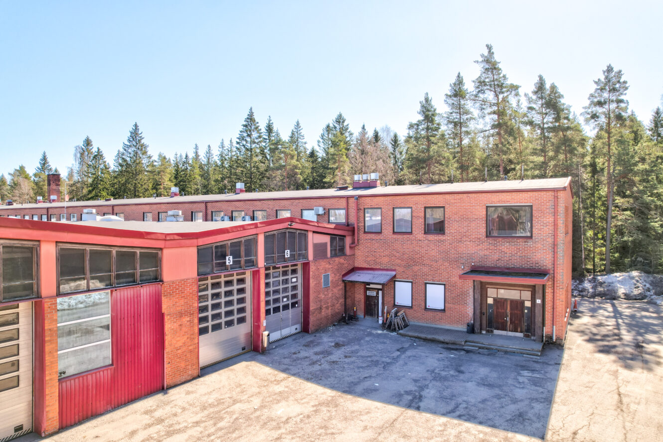 Tiilinen teollisuusrakennus, jossa on suuret autotallin ovet ja useita ikkunoita, puiden ympäröimänä kirkkaana päivänä.