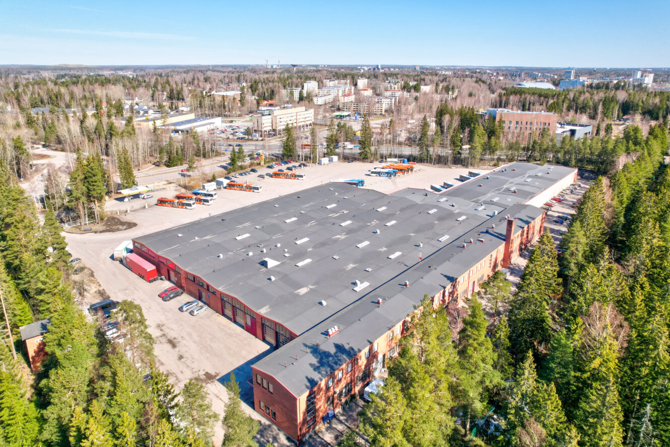 Suuri teollisuusrakennus, jossa on tasakatto ja viereinen parkkipaikka puiden ympäröimänä, ja taustalla näkyy kaupunkikuva.