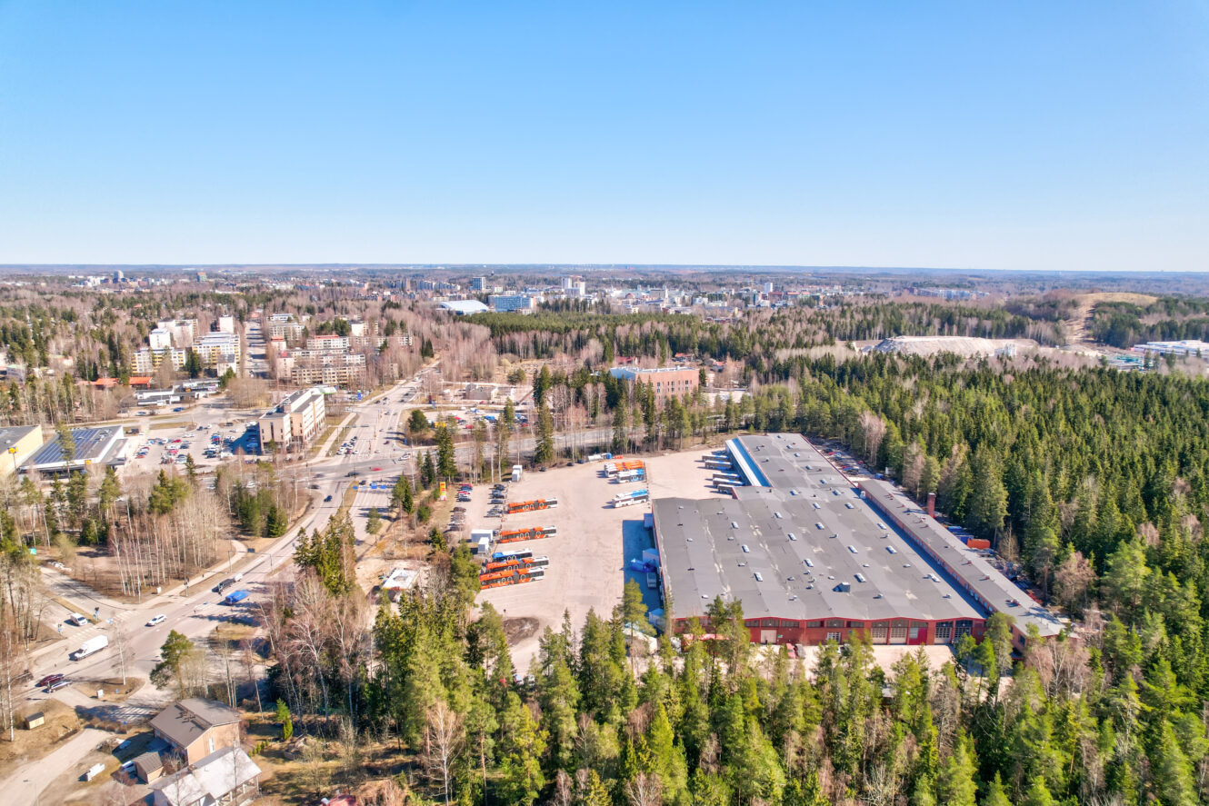Ilmakuva suuresta teollisuusvarastosta ja pysäköintialueesta, jota ympäröivät puut ja kaupunkirakennukset taustalla kirkkaalla taivaalla.