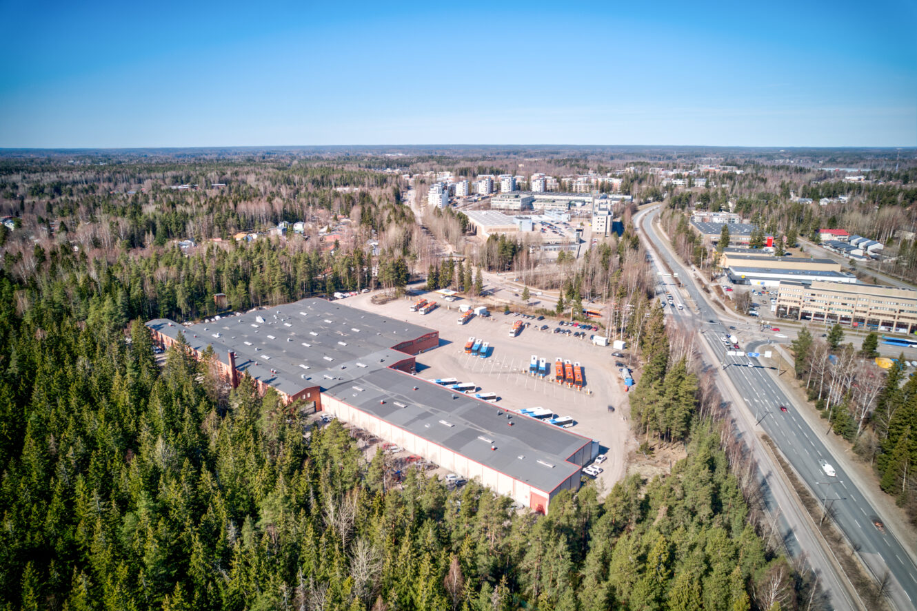 Ilmakuva puiden ympäröimästä suuresta varastosta, jonka taustalla näkyy päätietä ja rakennuksia kirkkaalla sinisellä taivaalla.