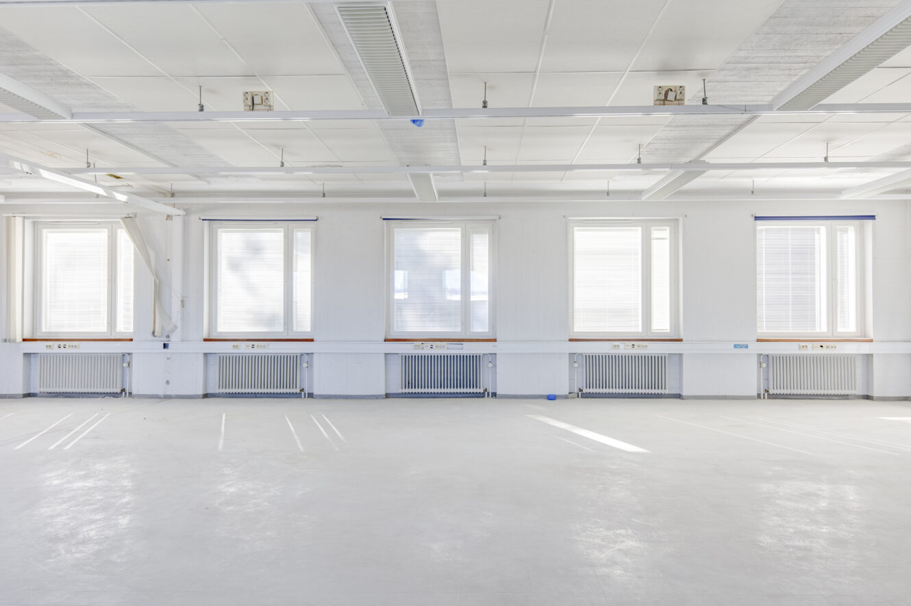 Tyhjä, valoisa huone, jossa on valkoiset seinät ja lattia, viisi suurta ikkunaa, joissa on kaihtimet, patterit jokaisen ikkunan alla ja kattovalaisimet yläpuolella.