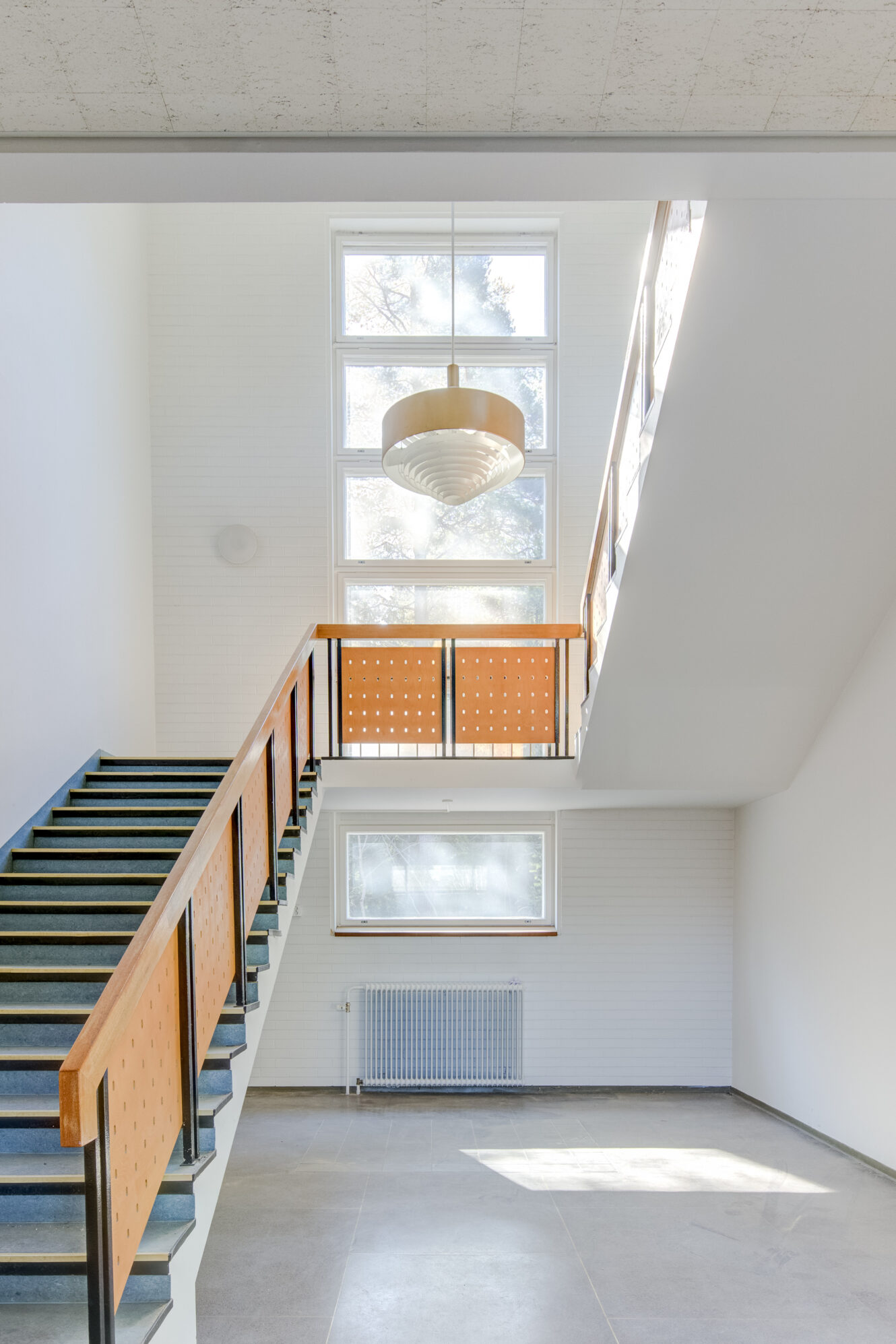 Moderni, minimalistinen portaikko, jossa on puiset kaiteet ja rei'itetyt paneelit suuren ikkunan vieressä, joka päästää luonnonvaloa valkoiseen sisätilaan.