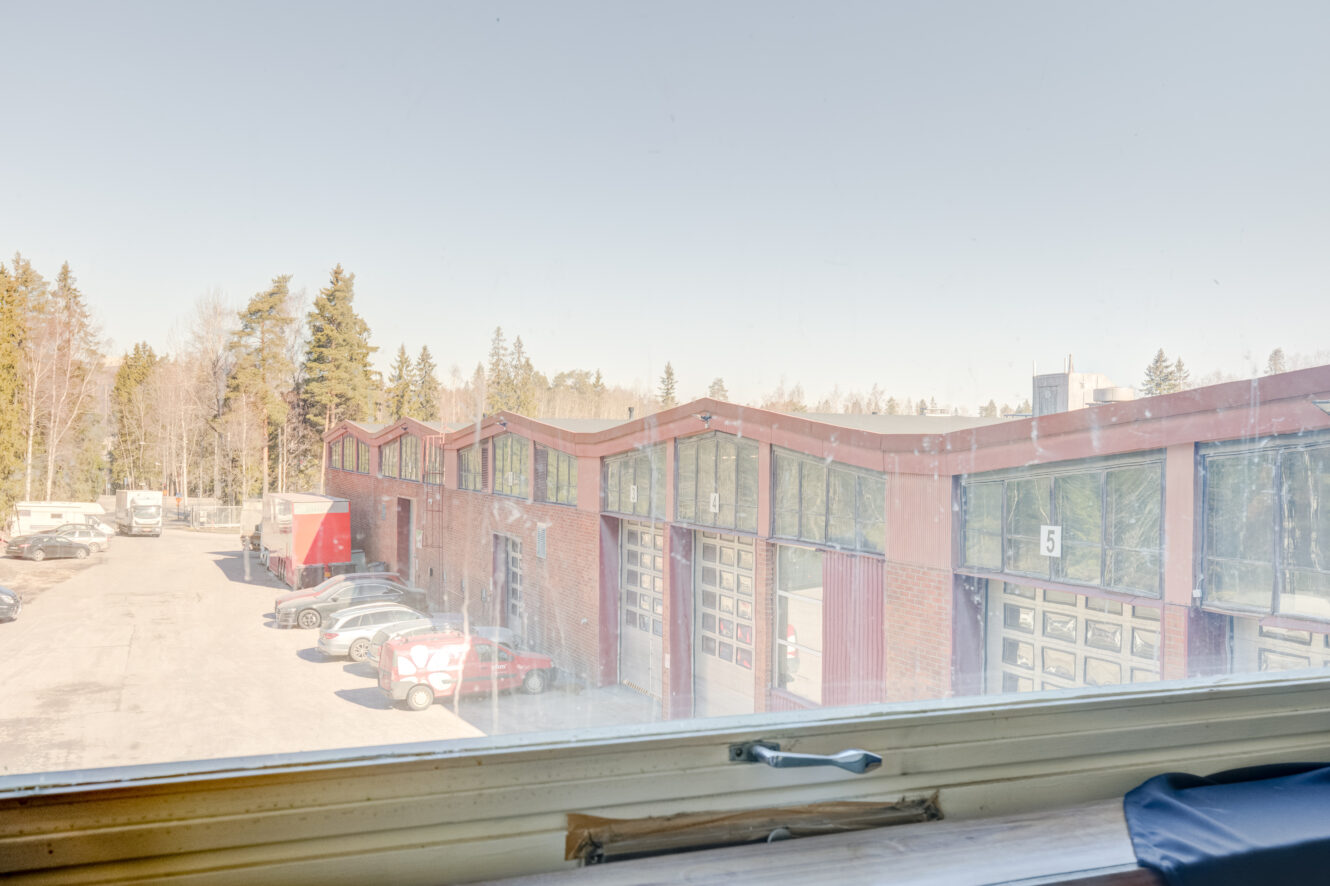 Näkymä ikkunasta, josta on näkymä punaisiin teollisuushallirakennuksiin, taustalla pysäköityjä autoja ja puita.