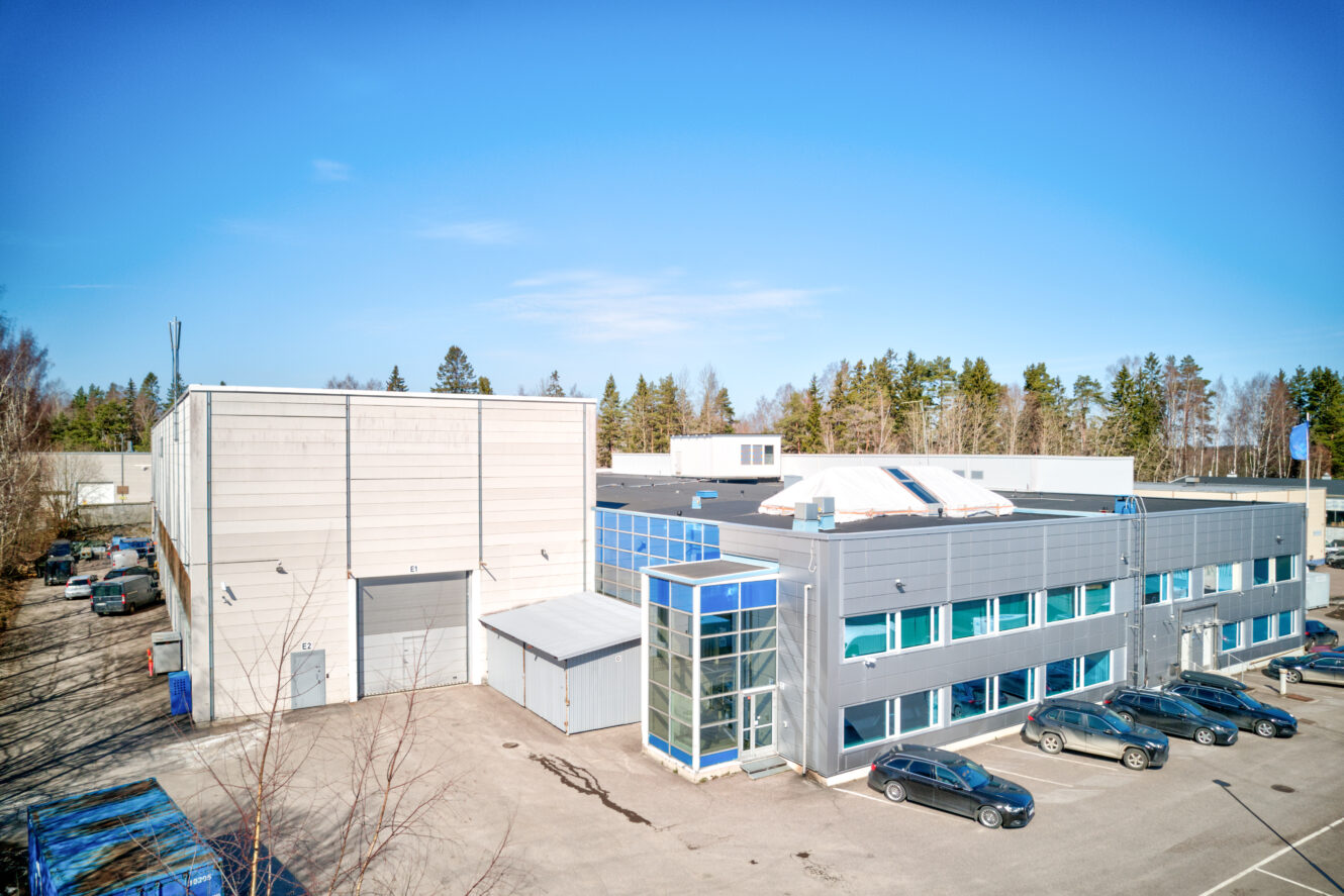 Moderni teollisuusrakennus, jossa on lasinen sisäänkäynti, useita pysäköityjä autoja ja kirkkaan sininen taivas; taustalla näkyy puita.