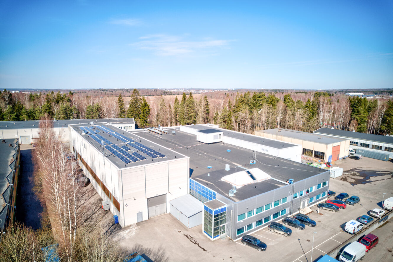 Ilmakuva modernista teollisuusrakennuksesta, jonka katolla on aurinkopaneeleita, pysäköityjen autojen, puiden ja muiden rakennusten ympäröimänä kirkkaan sinisen taivaan alla.