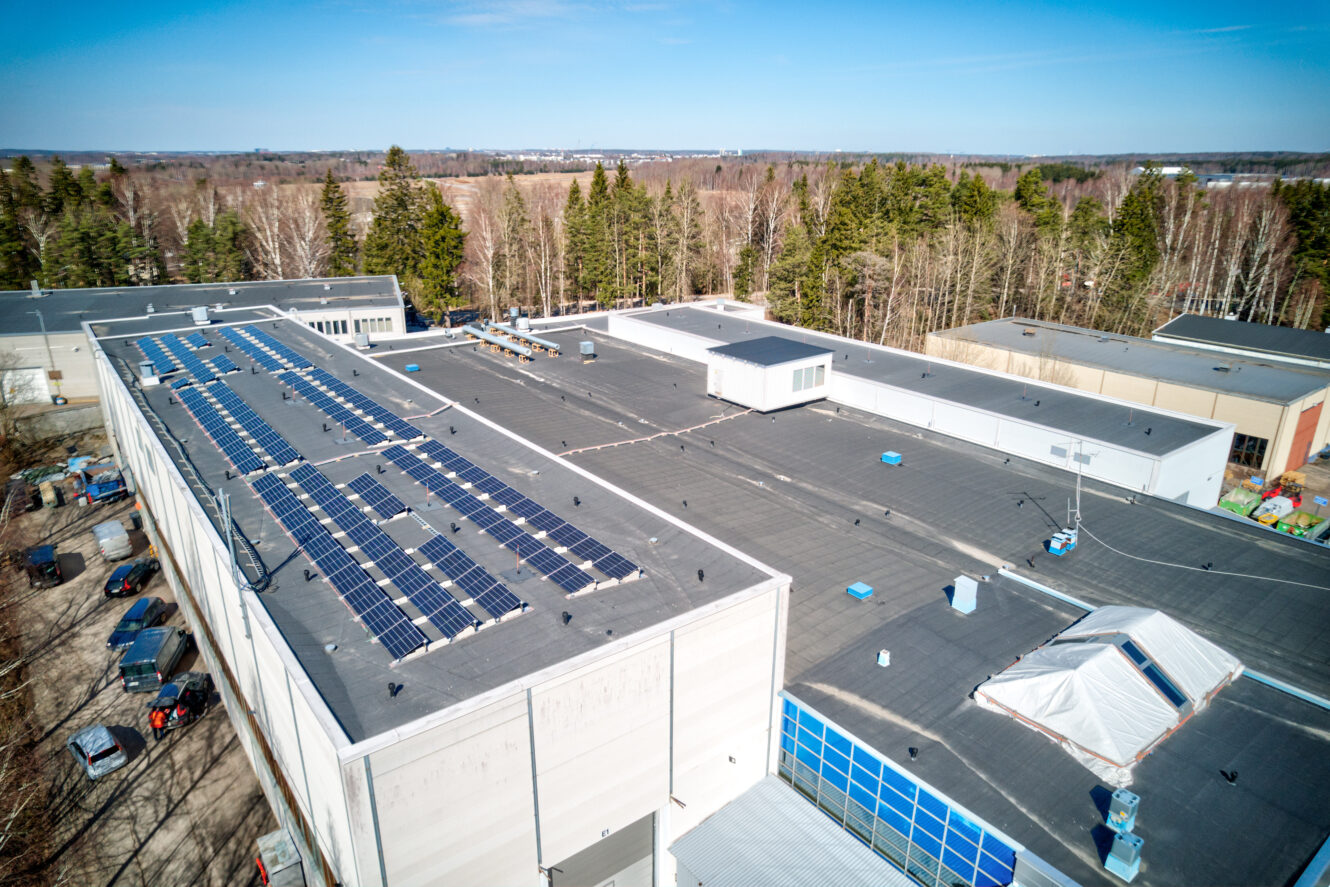 Ilmakuva tasakattoisesta teollisuusrakennuksesta, johon on asennettu aurinkopaneelit ja jota ympäröivät puut ja ulkovarastoalueet.