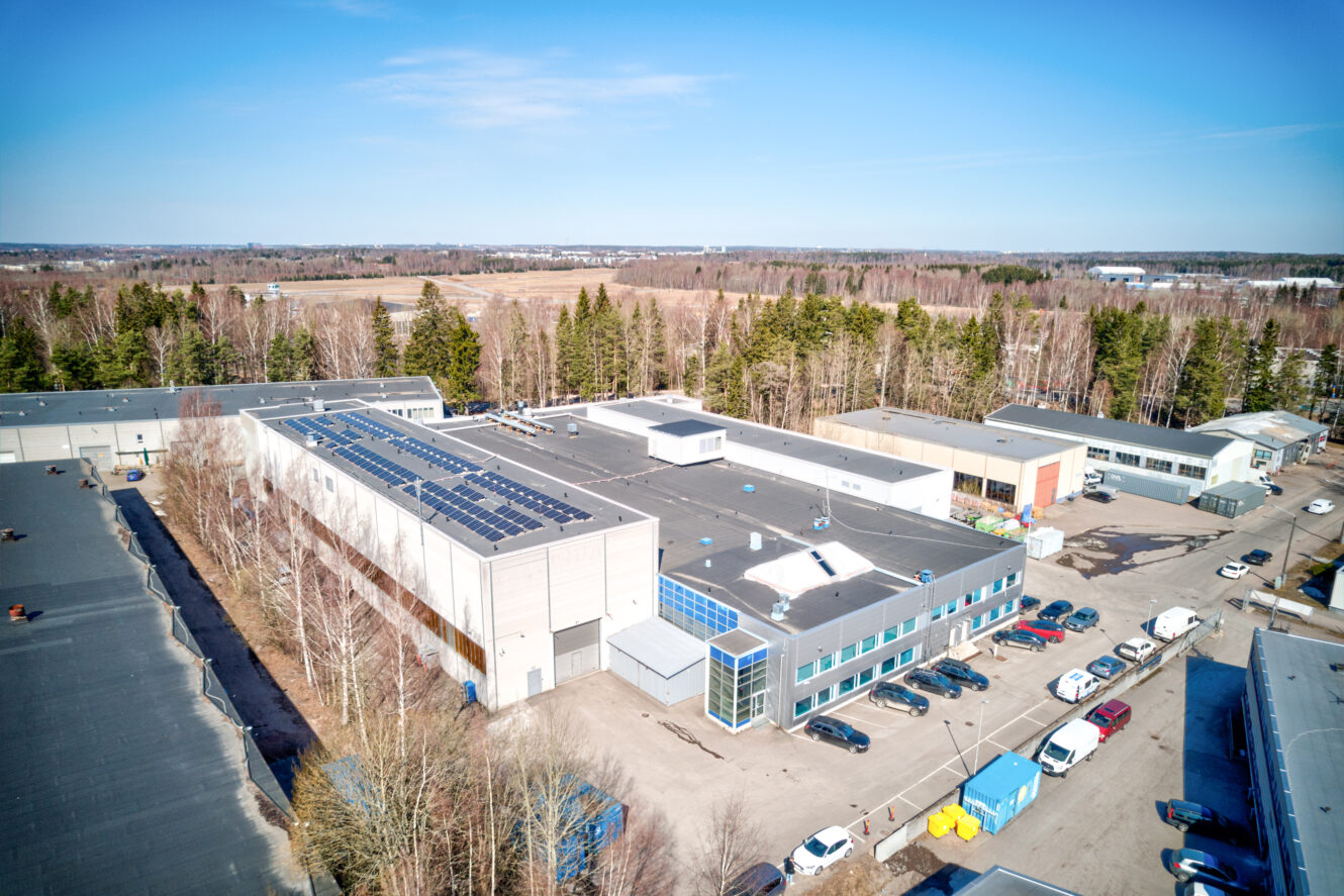 Ilmakuva teollisuusrakennuksesta, jonka katolla on aurinkopaneeleita, puiden, pysäköityjen ajoneuvojen ja viereisten rakenteiden ympäröimänä kirkkaan sinisen taivaan alla.