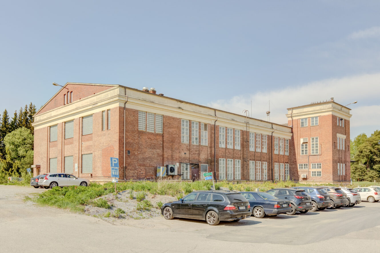 Suuri tiilinen teollisuusrakennus, jossa on paljon ikkunoita ja korkea sivurakennus, etualalla useita autoja pysäköitynä tontille.