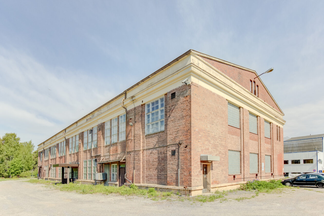 Suuri, kaksikerroksinen tiilinen teollisuusrakennus, jonka ikkunat on laudoitettu ja jota ympäröi tyhjä sorapelto ja harva kasvillisuus.