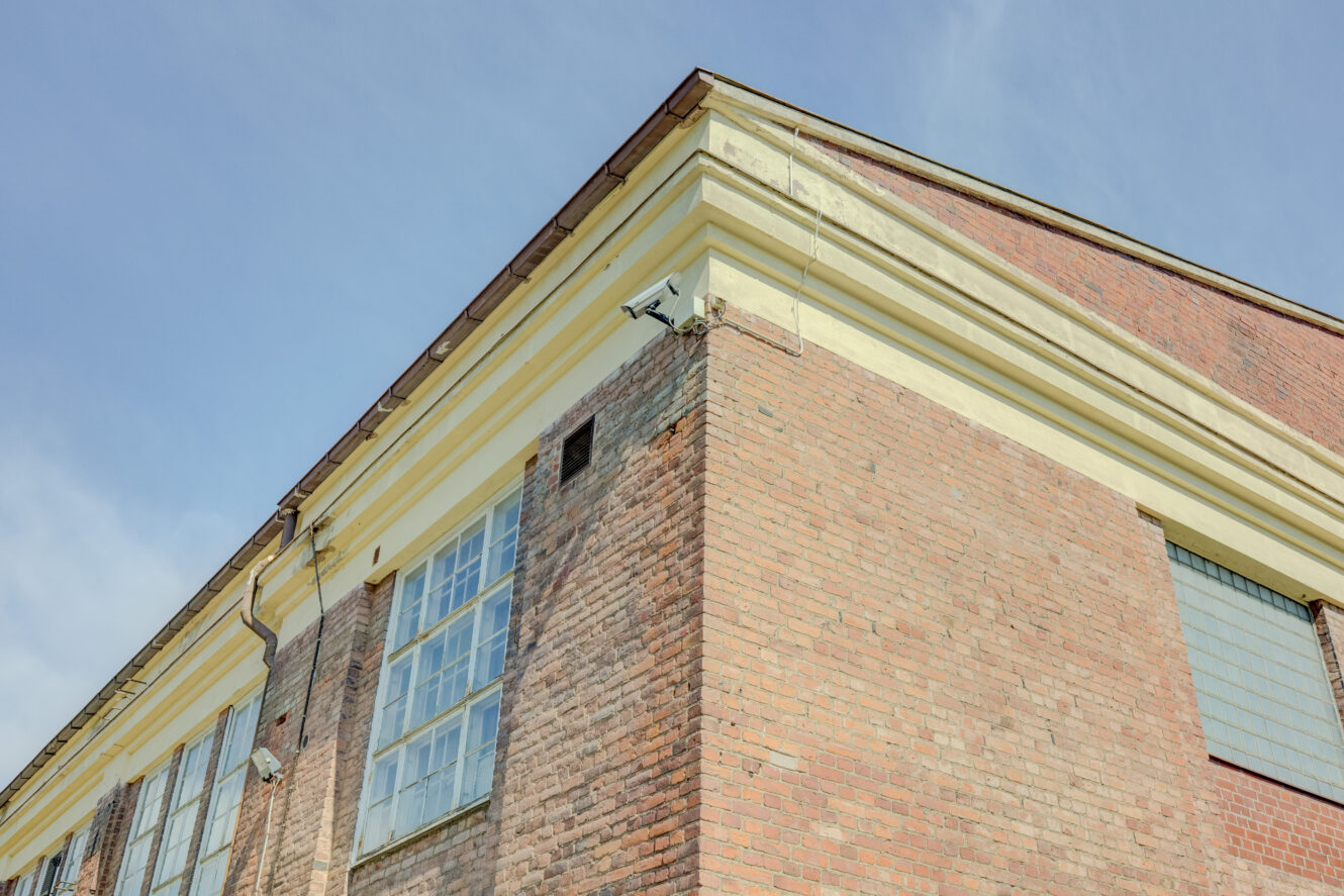 Valvontakamera, joka on asennettu tiilirakennuksen yläkulmaan, jossa on suuret ikkunat kirkkaalla sinisellä taivaalla.