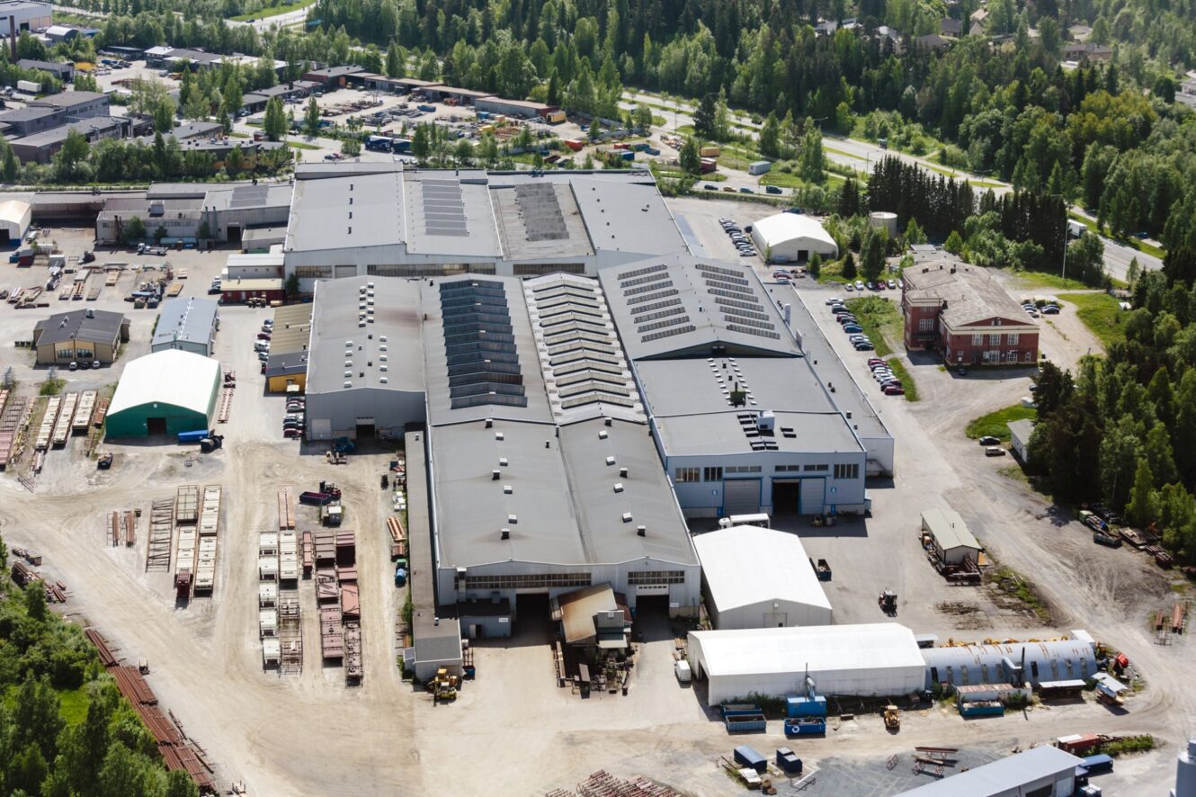 Ilmakuva suuresta teollisuuslaitoksesta, jossa on useita toisiinsa liittyviä varastorakennuksia, joita ympäröivät pysäköidyt ajoneuvot, varastointialueet ja puut.