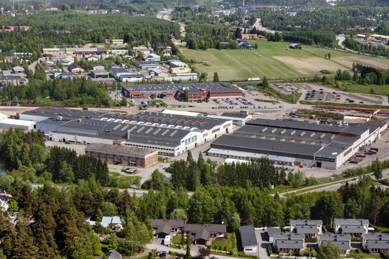 Ilmakuva teollisuusalueesta, jossa on suuria varastorakennuksia, pysäköintialueita ja ympäröivää viheraluetta, joka sijaitsee lähellä asuinalueita ja viljelysmaata.