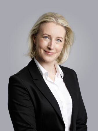 Vaaleatukkainen nainen, jolla on musta bleiseri ja valkoinen paita, hymyilee kameralle harmaata taustaa vasten.