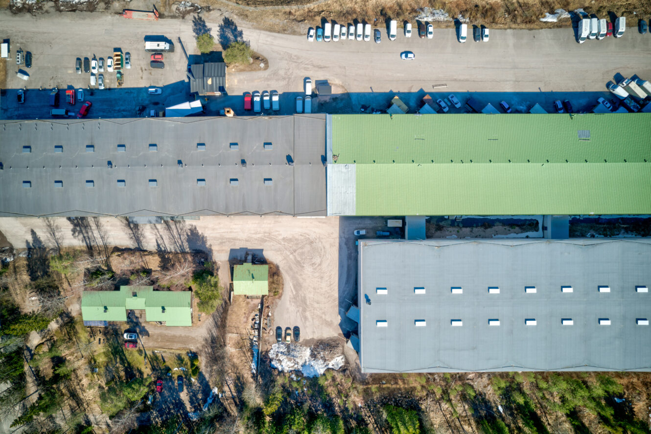 Ilmakuva teollisuusrakennuksista, joissa on harmaat ja vihreät katot ja joita ympäröivät pysäköidyt ajoneuvot ja hajallaan olevat puut likaisella tontilla.