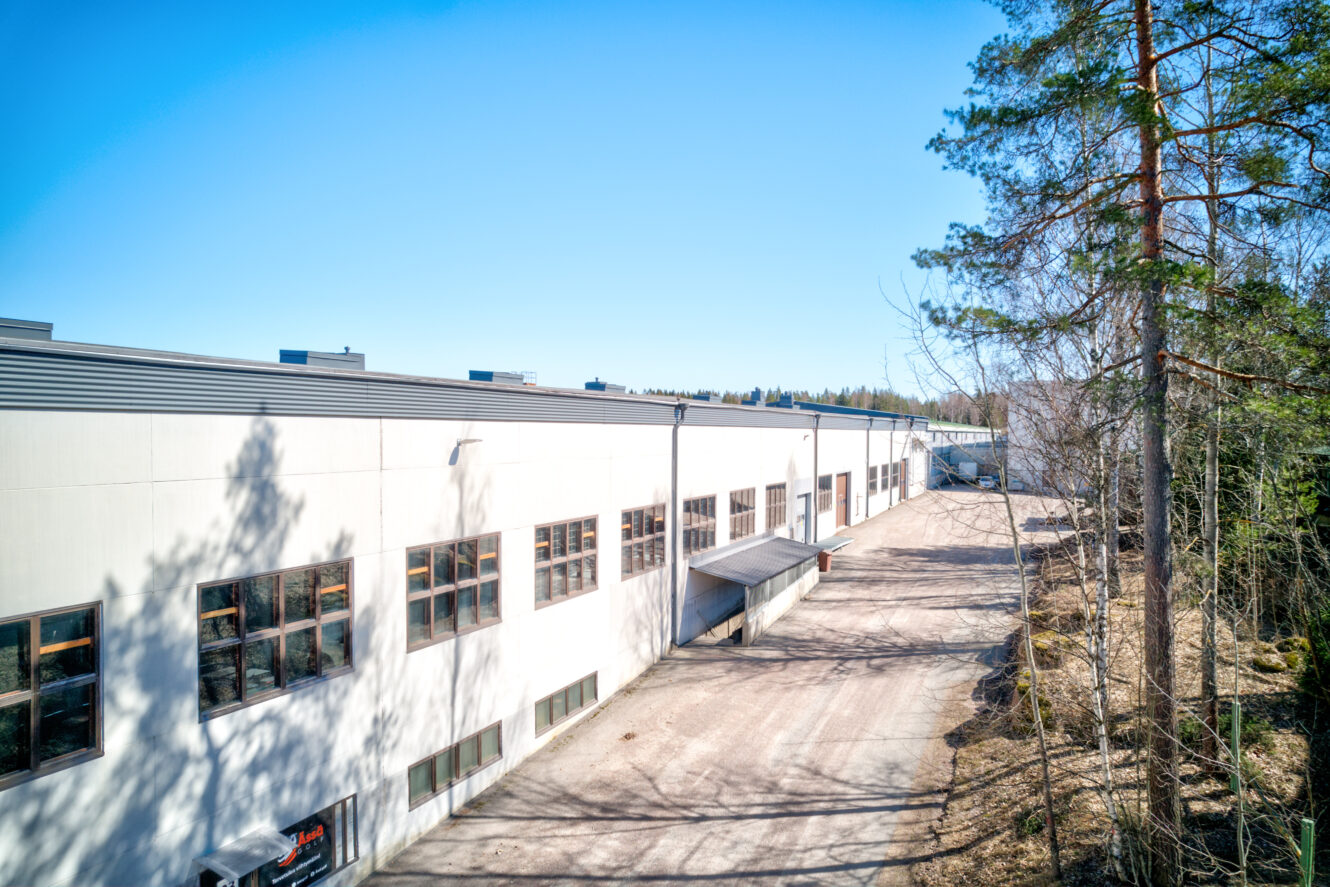 Suuri teollisuusvarastorakennus, jossa on useita ikkunoita, lastauslaituri ja päällystetty alue ulkona, jota reunustavat puut kirkkaana, aurinkoisena päivänä.