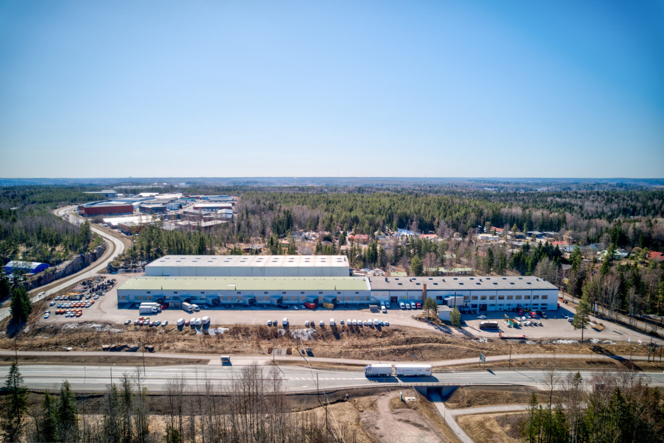 Ilmakuva teollisuusvarastokompleksista, jonka ulkopuolelle on pysäköity kuorma-autoja, metsän ja teiden ympäröimänä kirkkaalla sinisellä taivaalla.
