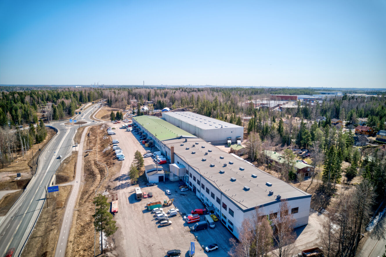 Ilmakuva teollisuuskompleksista, jossa on suuria varastoja, pysäköityjä ajoneuvoja ja läheisiä teitä metsäalueiden ympäröimänä kirkkaalla taivaalla.