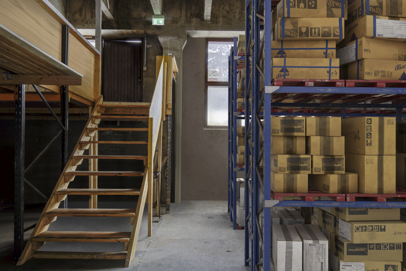 Varaston sisätila, jossa on pahvilaatikoita pinottuja metallihyllyjä, vasemmalla puuportaat ja taustalla ikkuna.