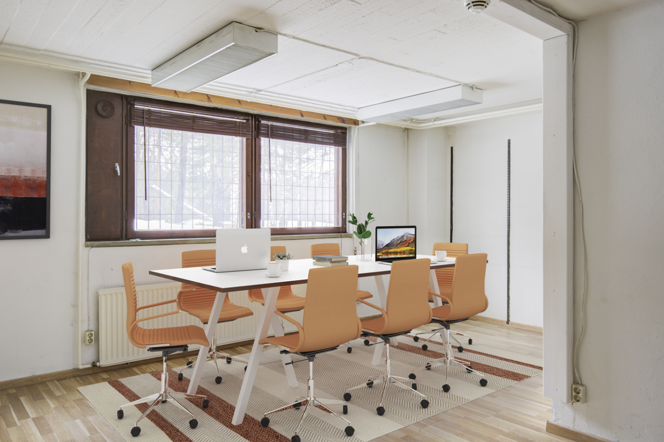 Moderni kokoushuone, jossa on valkoinen pöytä, kahdeksan oranssia tuolia, kaksi kannettavaa tietokonetta, kasvi ja kirjoja pöydällä. Näkyvissä on suuret ikkunat ja puulattia.