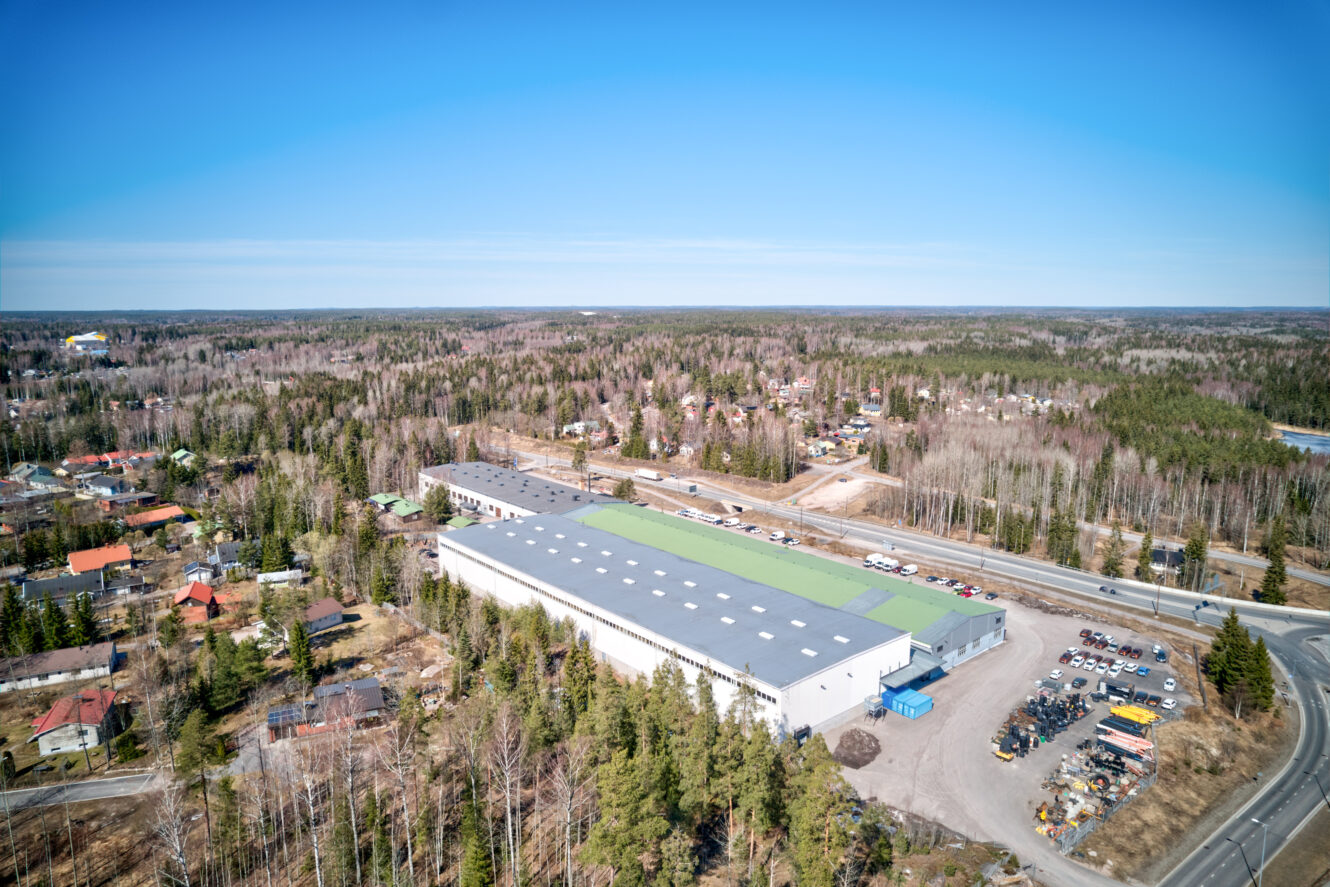 Ilmakuva puiden ympäröimästä suuresta teollisuusvarastosta, jonka lähellä on asuintaloja ja rakennuksen vieressä kulkee tie.