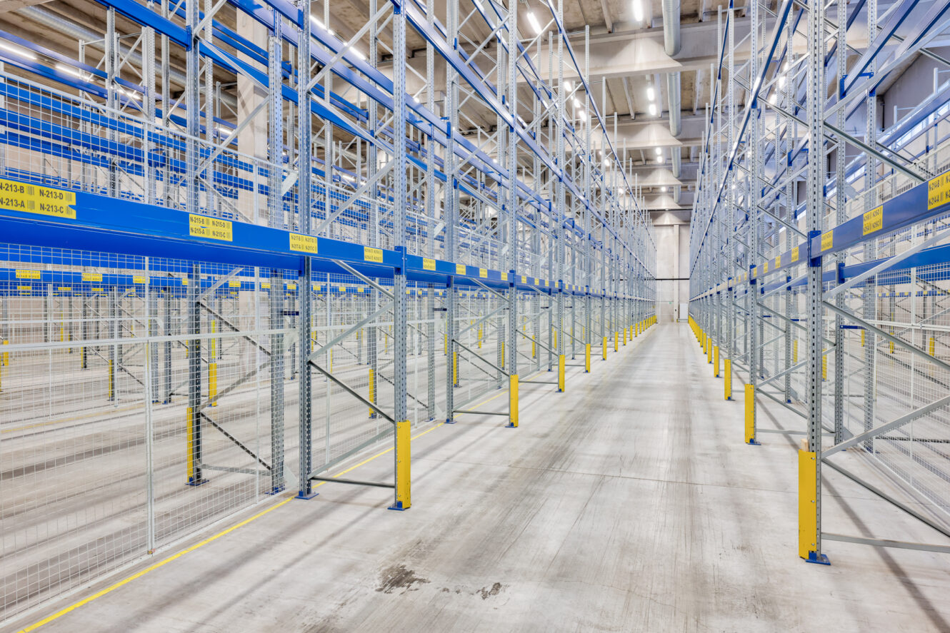 Tyhjä varaston sisätila, jossa on korkeita metallisia varastohyllyjä, betonilattia ja kirkas ylävalaistus.