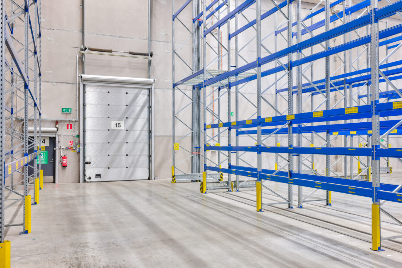 Tyhjä varasto, jossa on sinisiä metallihyllyjä, betonilattiat ja taustalla suuri suljettu teollisuusovi, jossa on merkintä 15. Vasemmalle seinälle on asennettu hätälaitteita.