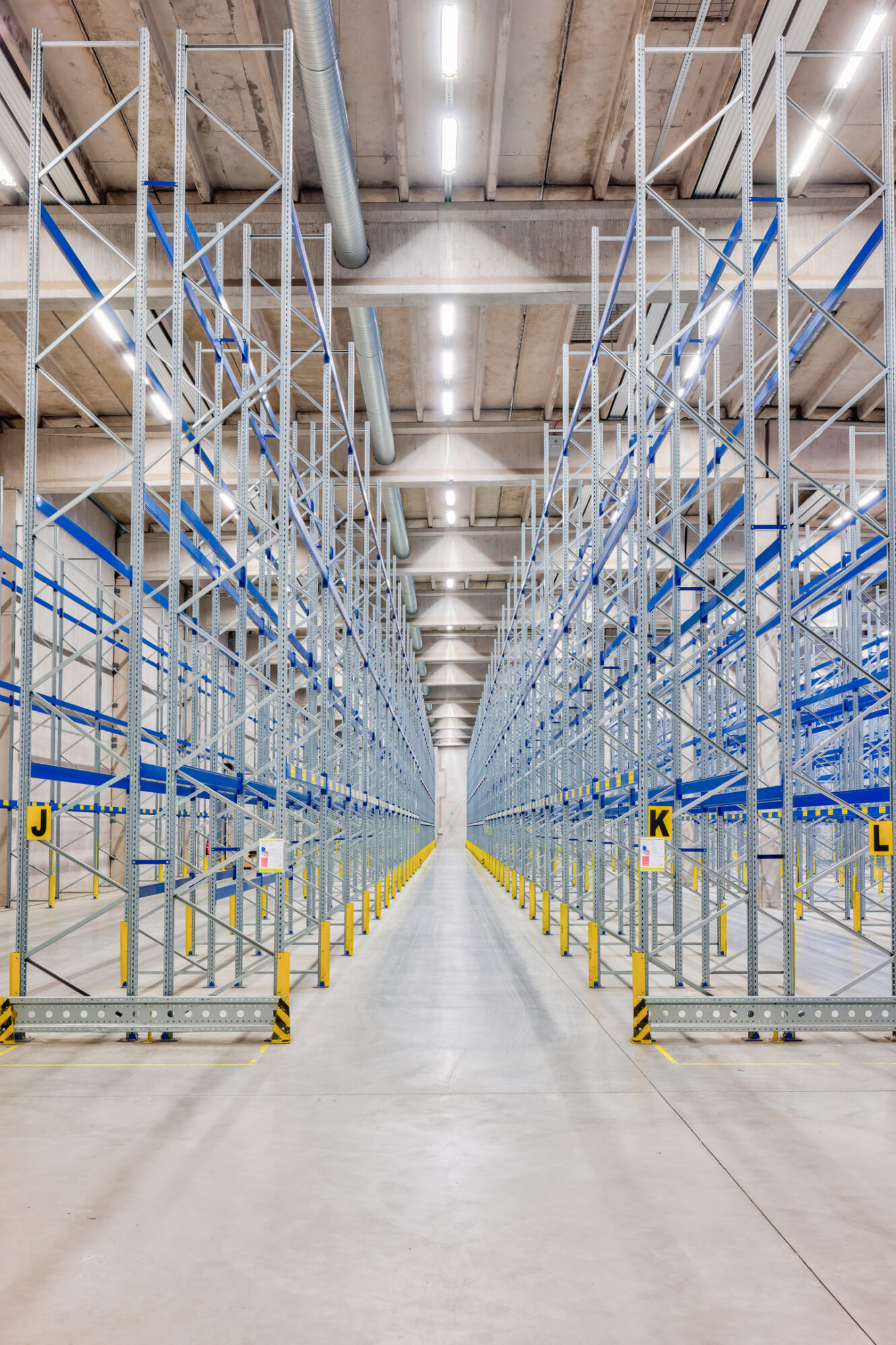 Tyhjä teollisuusvarasto, jossa on rivejä korkeita metallisia varastohyllyjä, jotka on merkitty kirjaimilla, kirkkaiden kattovalaisimien ja betonilattioiden alla.