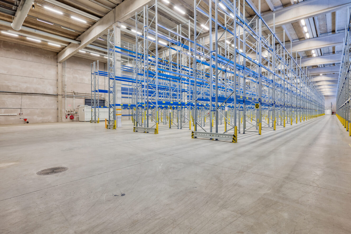 Tyhjä teollisuusvaraston sisätila, jossa on korkeita metallisia varastohyllyjä, betonilattia ja loisteputkivalaisimet.
