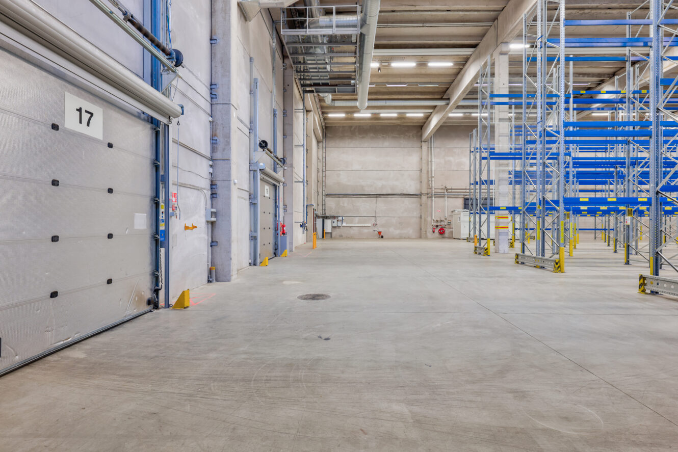 Tyhjä teollisuusvaraston sisätila, jossa on betonilattia, lastauslaiturin ovet vasemmalla ja siniset metalliset varastohyllyt oikealla.