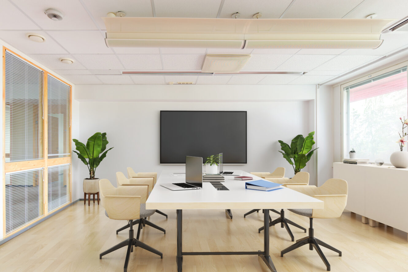 Moderni kokoushuone, jossa on suuri näyttö, valkoinen pöytä, beiget tuolit, kannettavat tietokoneet, muistikirjat, kaksi ruukkukasvia ja suuret ikkunat, jotka päästävät luonnonvaloa sisään.