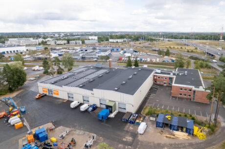 Ilmakuva suuresta teollisuusvarastosta, jossa on lastauslaitureita, pysäköityjä ajoneuvoja ja ympäröiviä toimistorakennuksia kaupallisella alueella.