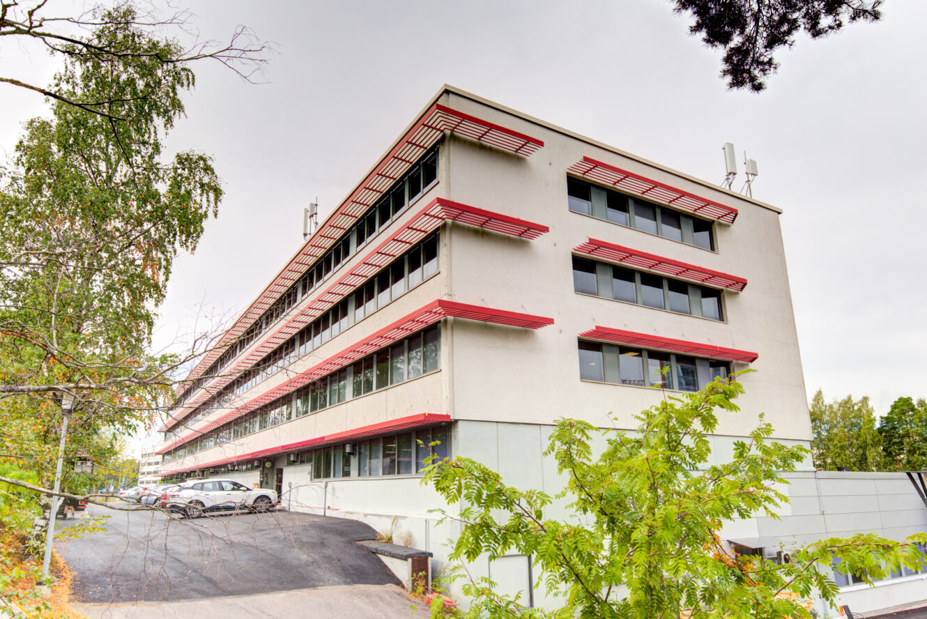 Moderni, monikerroksinen toimistorakennus, jossa on punaisia korostuksia ja suuret ikkunat, puiden ja pysäköityjen autojen ympäröimänä pilvisenä päivänä.