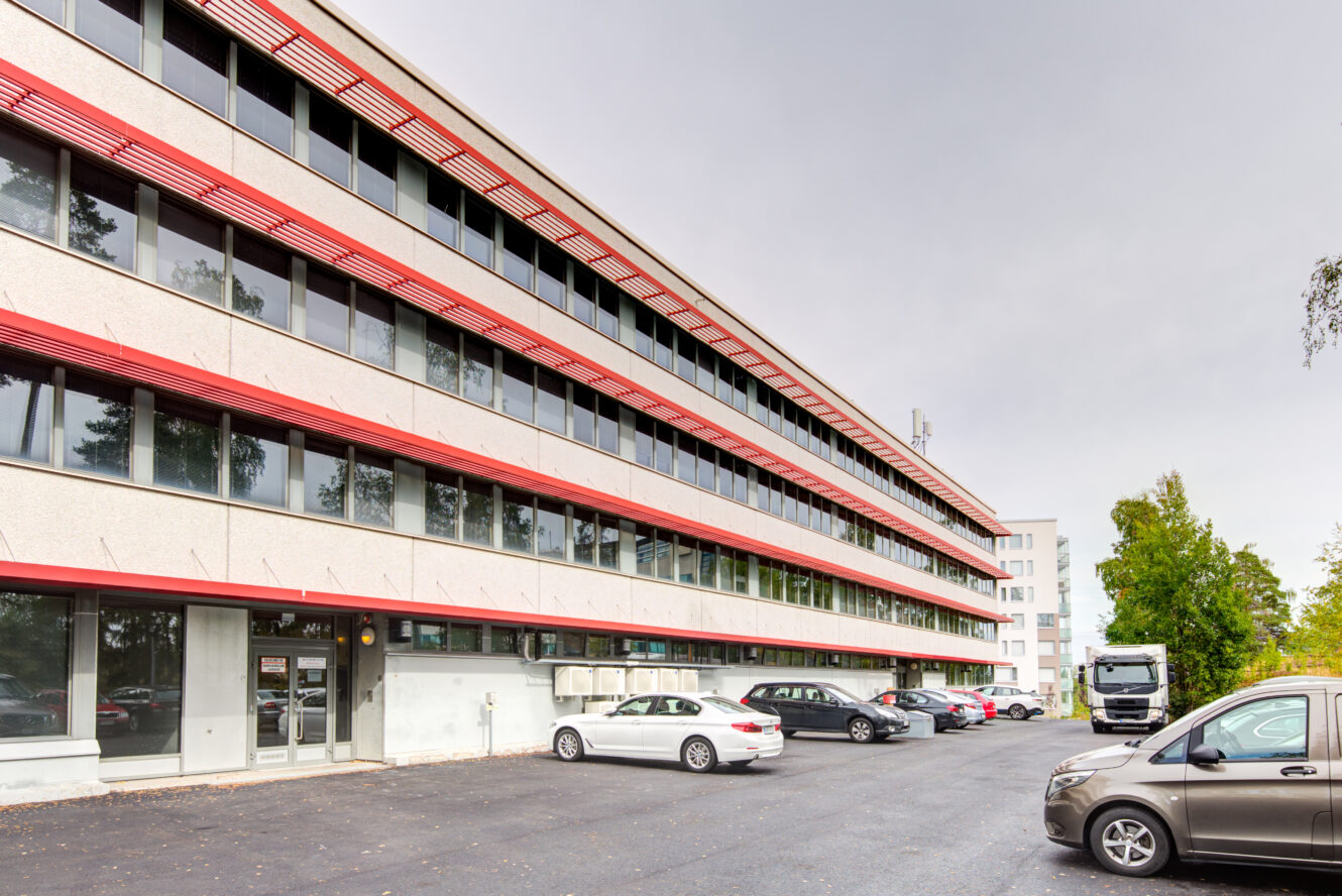 Nelikerroksinen toimistorakennus, jossa on punaiset korostukset, suuret ikkunat ja useita autoja pysäköitynä viereiselle parkkipaikalle pilvisenä päivänä.