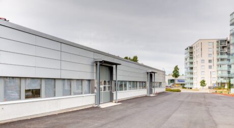 Pitkä, moderni teollisuusrakennus, jossa on harmaita paneeleita ja suuret ikkunat, asfaltoidun ajoväylän vieressä, taustalla näkyvät kerrostalot.