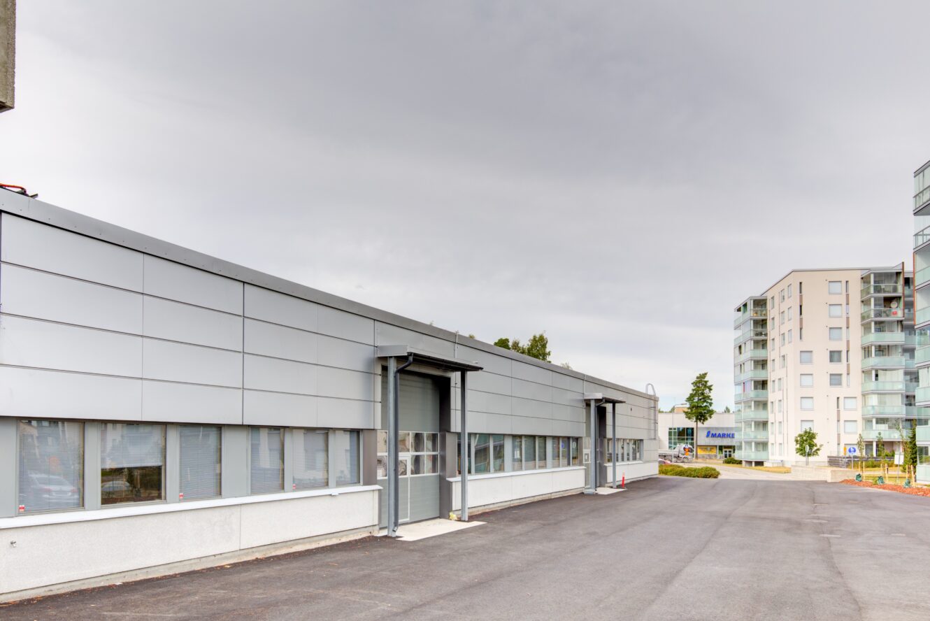 Nykyaikainen teollisuusrakennus, jossa on metalliverhous ja suuret ikkunat, päällystetyn pysäköintialueen vieressä, taustalla näkyvät kerrostalot pilvisen taivaan alla.