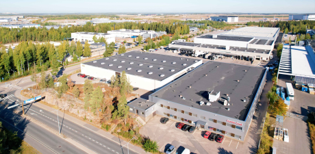 Ilmakuva teollisuusalueesta, jossa on suuria varastoja, toimistorakennuksia, ympäröiviä puita, pysäköityjä ajoneuvoja ja läheisiä teitä päivällä.