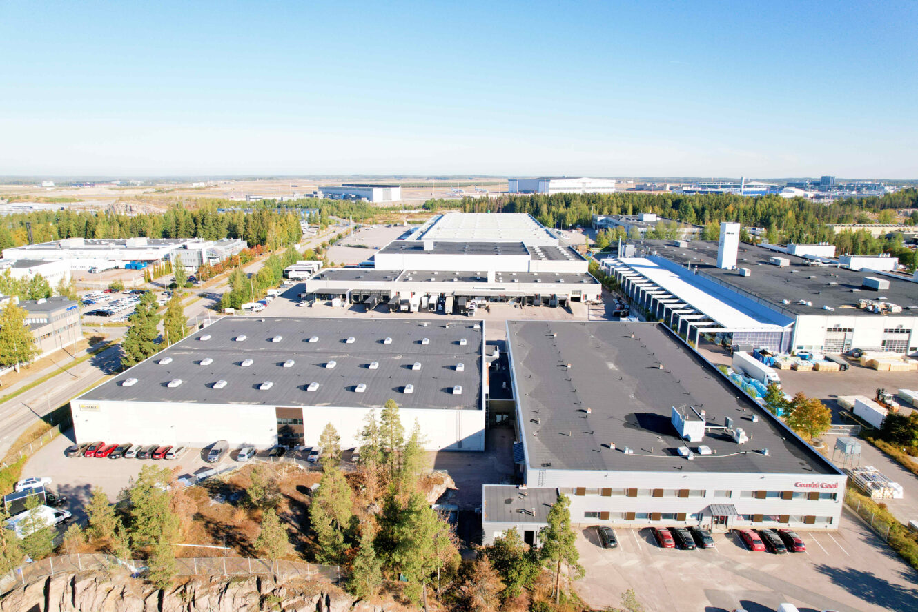 Ilmakuva suuresta teollisuusalueesta, jossa on useita rakennuksia, pysäköintialueita ja ympäröiviä puita kirkkaalla taivaalla.