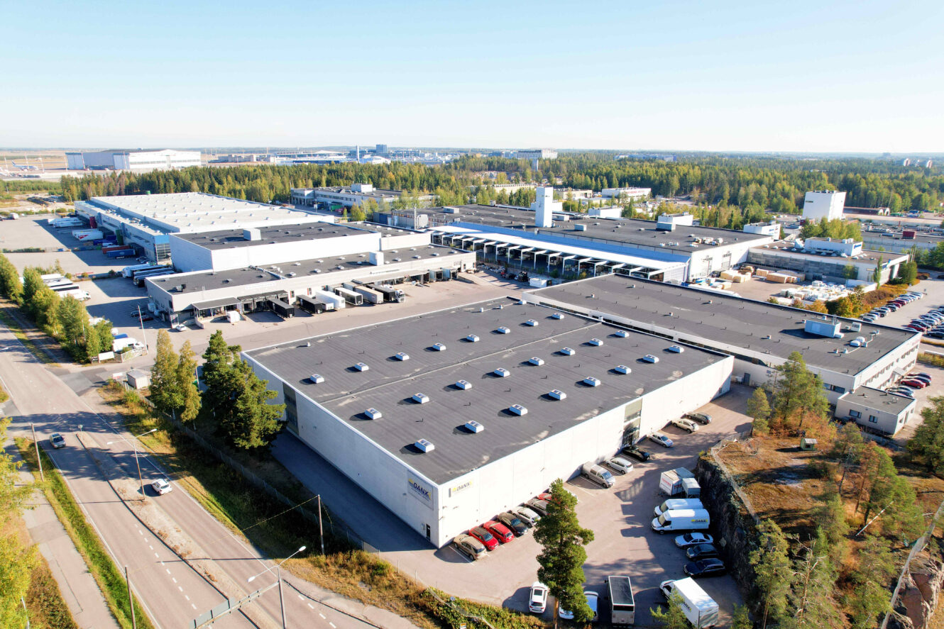 Ilmakuva suuresta teollisuuskompleksista, jossa on useita varastorakennuksia, lastauslaitureita, pysäköityjä autoja ja ympäröivää viheraluetta.