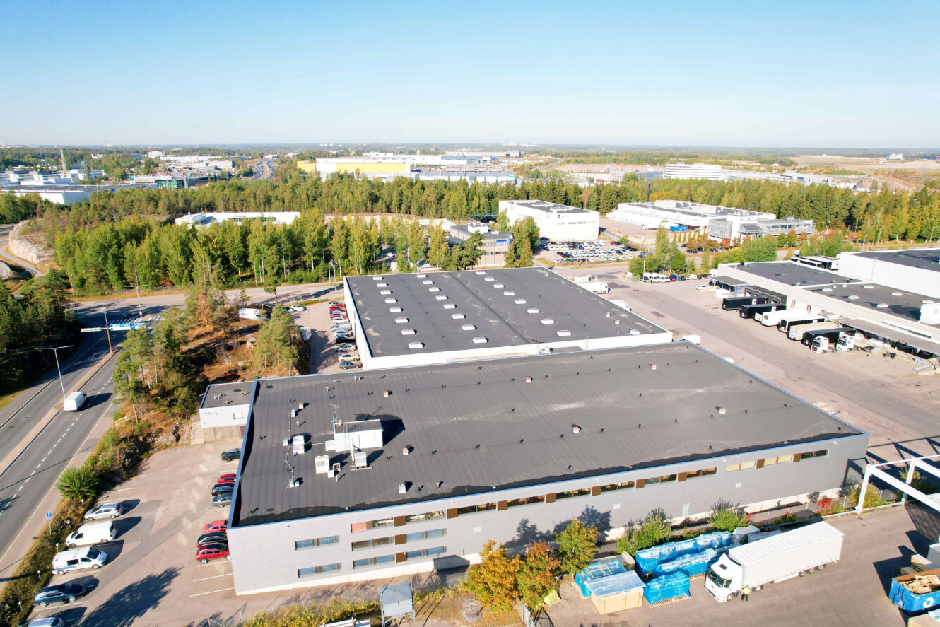 Suurta tasakattoista teollisuusrakennusta ympäröivät pysäköidyt ajoneuvot, puut ja muut liikerakennukset kirkkaalla taivaalla.