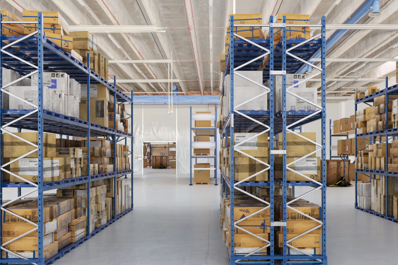 Varaston sisätila, jossa on sinisiä metallihyllyjä täynnä pahvilaatikoita ja paketteja, jotka on järjestetty riveihin puhtaalle, vaaleanharmaalle lattialle.
